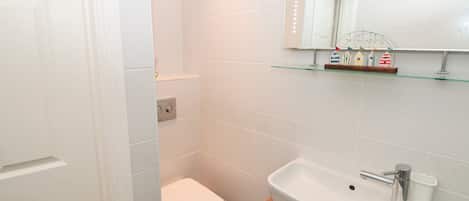 A bathroom with white tiles, a white toilet, a sink with a mirror above it, and a shelf with decorative items.