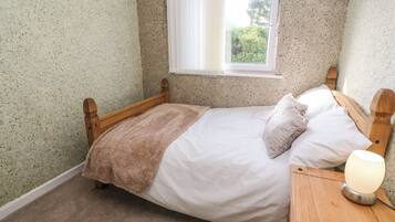 A bedroom with a wooden bed, white bedding, a brown throw blanket, a bedside lamp, and a window with a view of greenery.