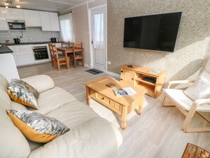 A modern living room with a sofa, coffee table, and wooden side table, a TV mounted on the wall, and a kitchen in the background. - Valhalla (St Ives)