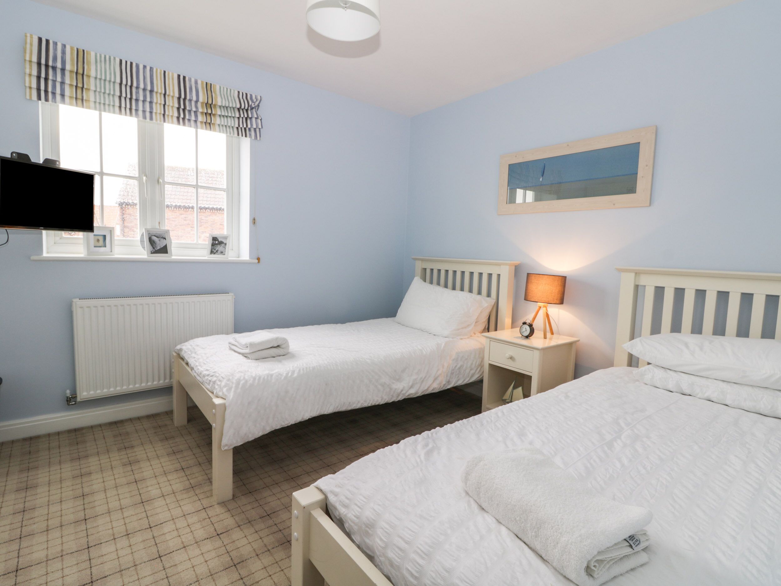 A bedroom with two beds, a radiator, a window with curtains, a bedside table with a lamp, and a framed picture on the wall.