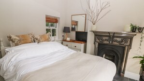 A bedroom with a bed, nightstand, dresser, mirror, fireplace, and a window with a view of greenery. - Yew Tree Cottage (York)