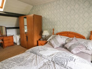 A bedroom with a wooden wardrobe, a bedside table with a lamp, a bed with patterned bedding, and a mirror on the dresser. - Caerau Farm House (Caersws)