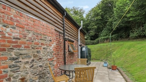 A patio with a round table and two wooden chairs, a brick wall, and a green garden area. - Caerau Farm House (Caersws)