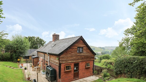 A rustic wooden house with a brick extension, a small garden, and a view of rolling hills in the background. - Caerau Farm House (Caersws)