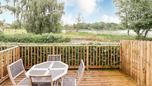 Une terrasse en bois, agrémentée d’une table et de chaises, entourée d’une clôture en bois, et offrant une vue sur une rivière et la verdure environnante.