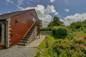 Une maison rustique en bois, avec des fondations en pierre, un escalier en bois et un jardin bien entretenu où poussent diverses plantes et fleurs.