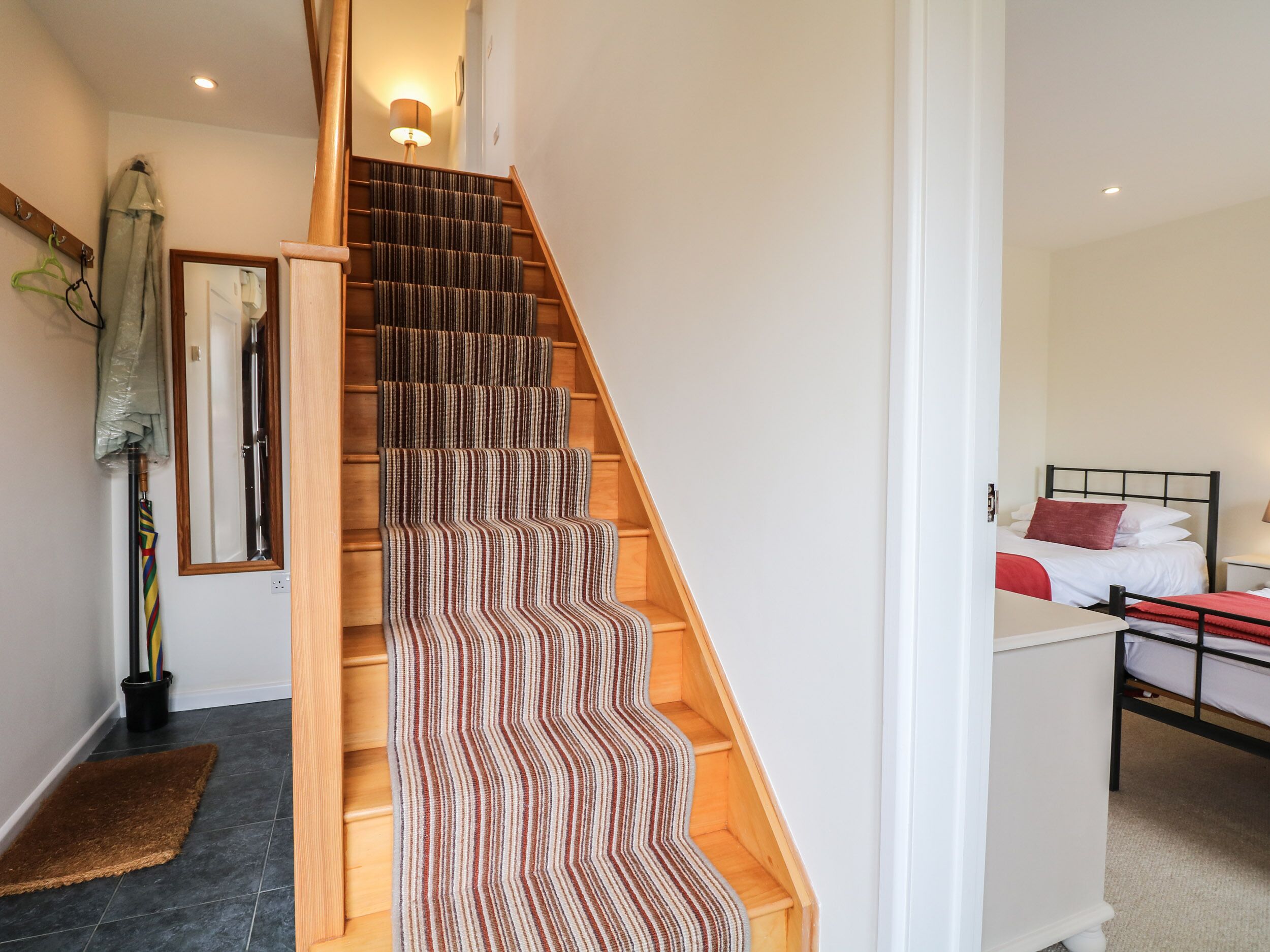 A staircase with striped carpet, a wooden handrail, a mirror, and a bed with a red pillow in the adjacent room.
