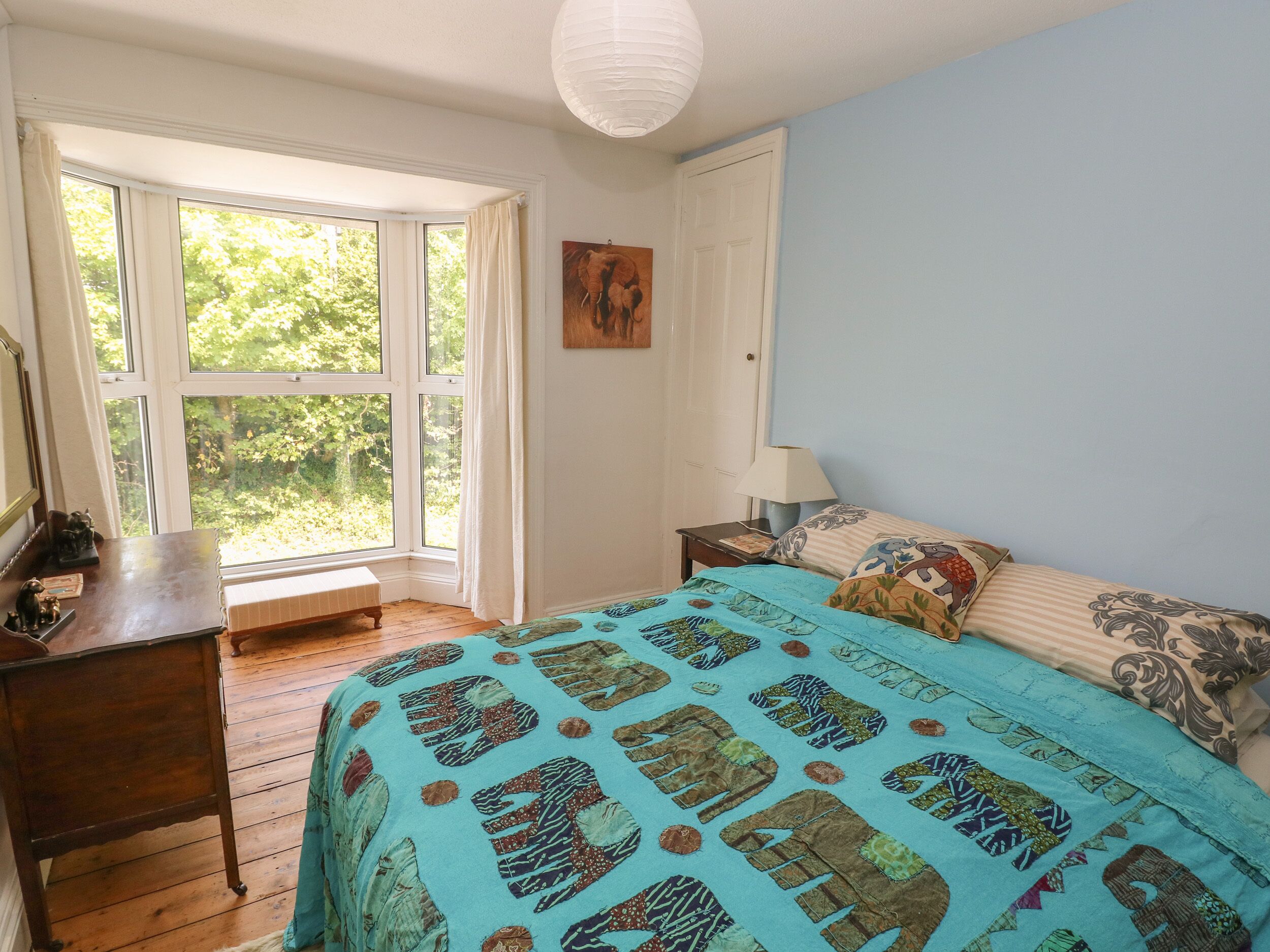 Quarto com uma grande janela, uma cama com roupa de cama estampada, uma mesinha de cabeceira com abajur e um pequeno banco perto da janela.
