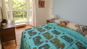 A bedroom with a large window, a bed with patterned bedding, a bedside table with a lamp, and a small bench near the window. - Hawthorn House (Pembroke)