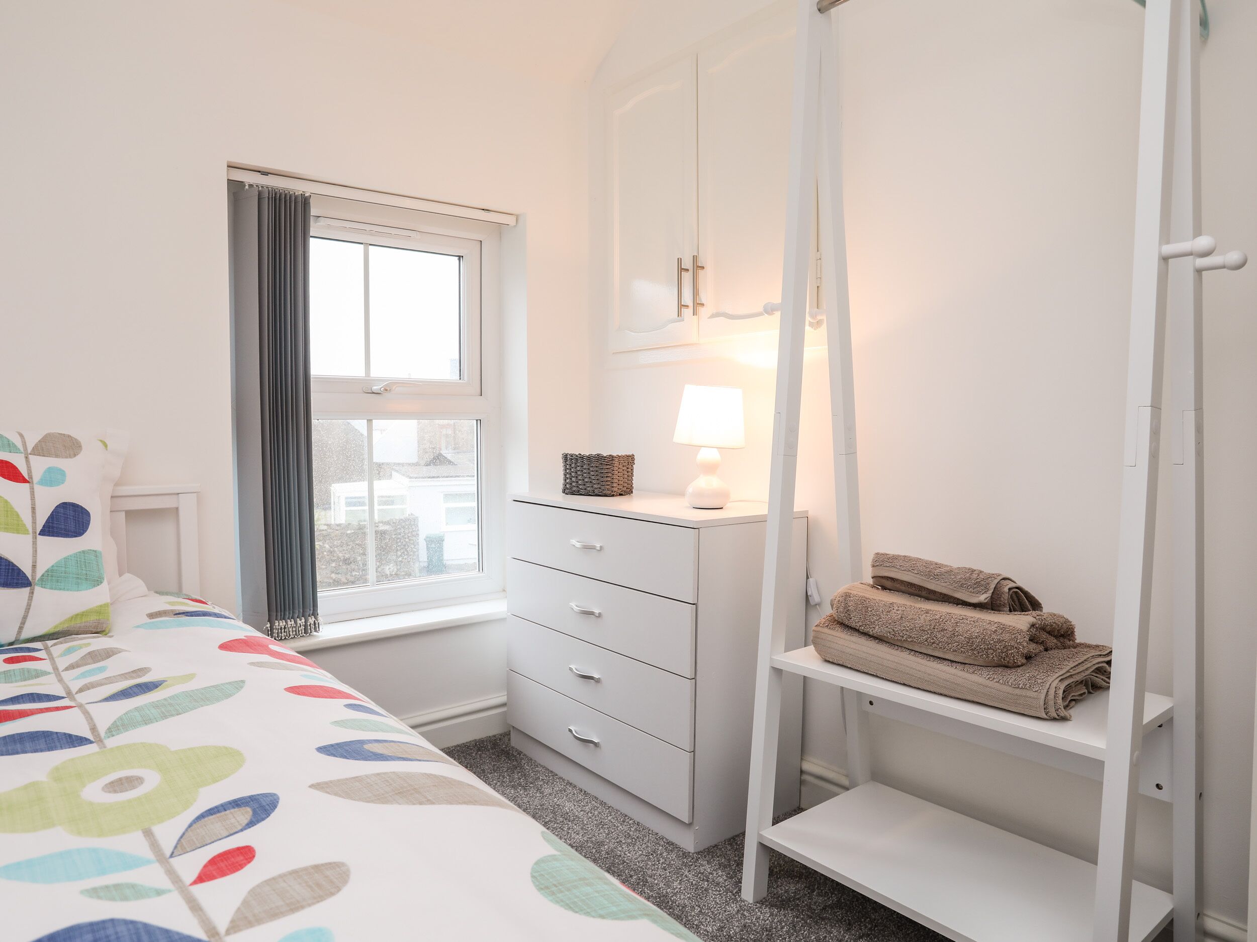 Une chambre à coucher comprenant un lit, une commode, une étagère avec des serviettes, une fenêtre avec des rideaux et une lampe de chevet.