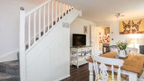 A staircase with white railings leading to a room with a television, a dining table with chairs, and a kitchen area visible through an open door. - Bwthyn Defaid (Pwllheli)