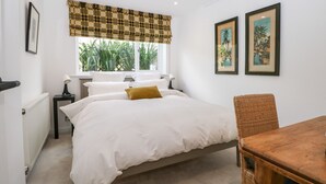 Un dormitorio con cama, ventana con cortinas, mesita de noche, una silla y dos cuadros en la pared.
