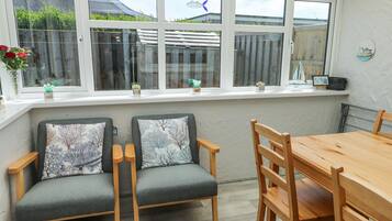 A dining area with a wooden table and chairs, a window with a view of a garden and a building, and a shelf with decorative items.