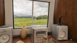 Ein Radiosystem, eine Vase und Lautsprecher auf einem Holztisch neben einem Fenster mit Blick auf ein Feld und Berge.