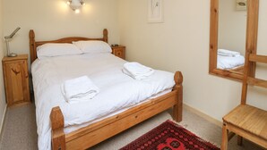 Ein ordentlich bezogenes Bett mit weißen Leinen, ein Holzkopfteil, ein Nachttisch mit Lampe, ein Teppich mit rotem Muster und ein großer Spiegel an der Wand.