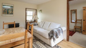 Ein gemütliches Wohnzimmer mit einem weißen Sofa, einem Holzespeisetisch und Stühlen. Ein Fenster mit weißem Vorhang, eine Lampe und ein Gemälde sind an der Wand zu sehen.