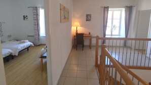 Une pièce avec un plancher en bois, un bureau avec une lampe, un lit recouvert d’une courtepointe blanche et une fenêtre avec des rideaux.