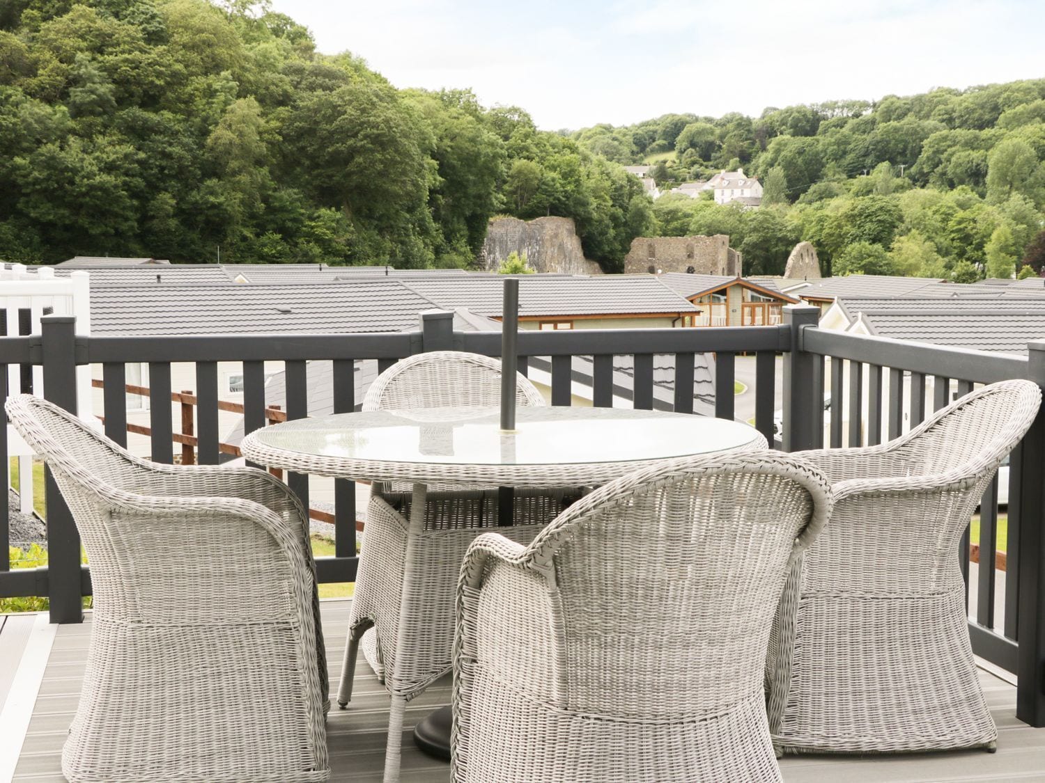 A round table with four wicker chairs on a deck with a black railing, surrounded by lush greenery and a distant view of buildings.