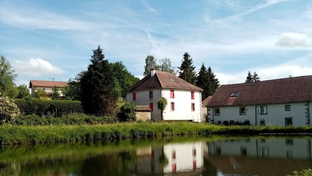 Une scène paisible au bord de la rivière, avec des bâtiments traditionnels, une étendue d’eau calme qui reflète les structures et une végétation luxuriante.