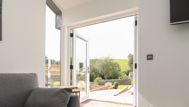 A modern living room with a grey sofa, a wooden table, and a large sliding glass door leading to an outdoor patio. - Ghyll Cottage (Milnthorpe)
