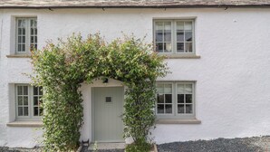A white building with a grey door, two windows on the upper floor, and a small porch area with greenery. - Ghyll Cottage (Milnthorpe)