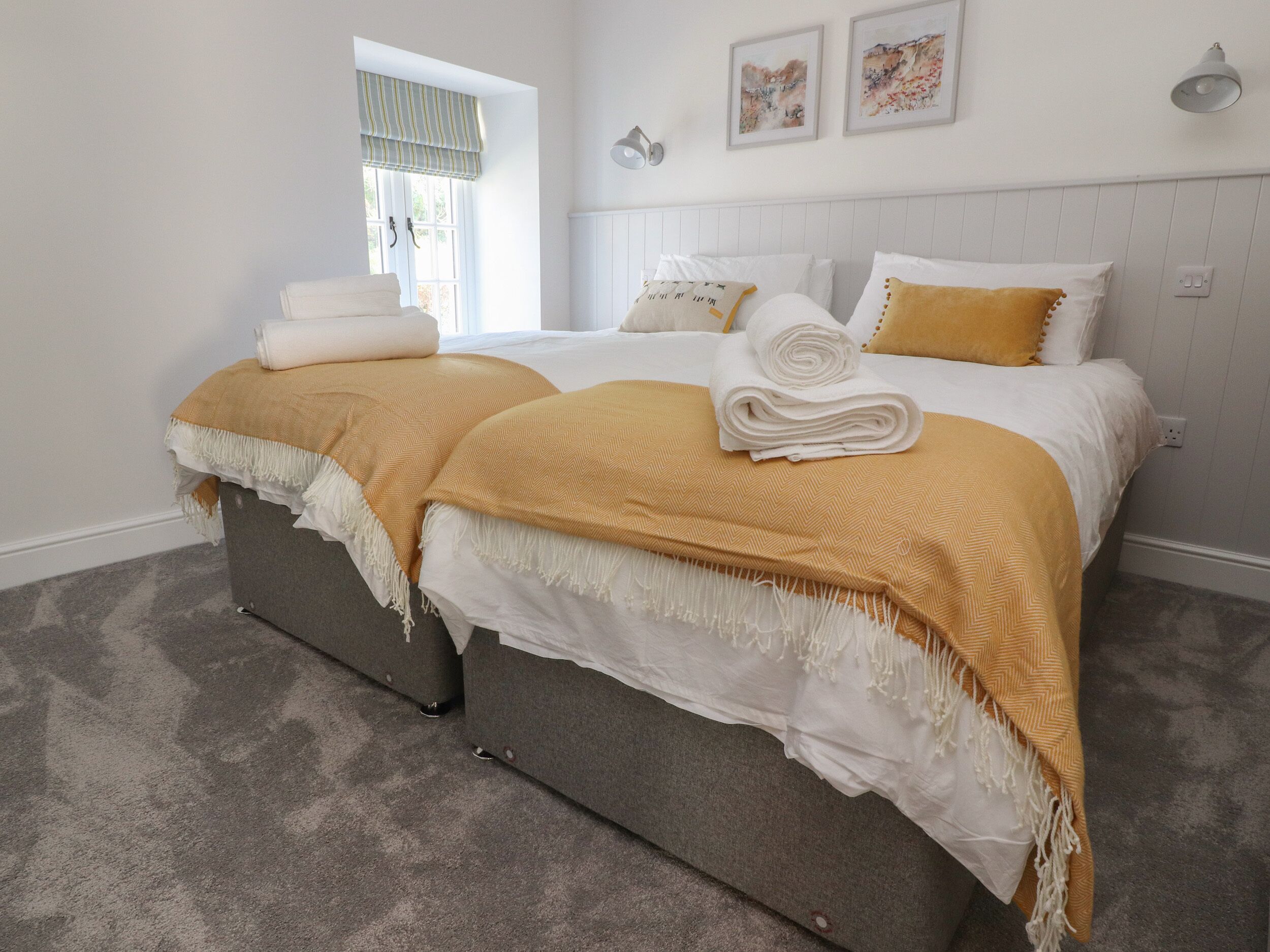 Una cama doble con ropa de cama blanca y mostaza, una ventana con cortina a rayas y dos lámparas fijadas a la pared.