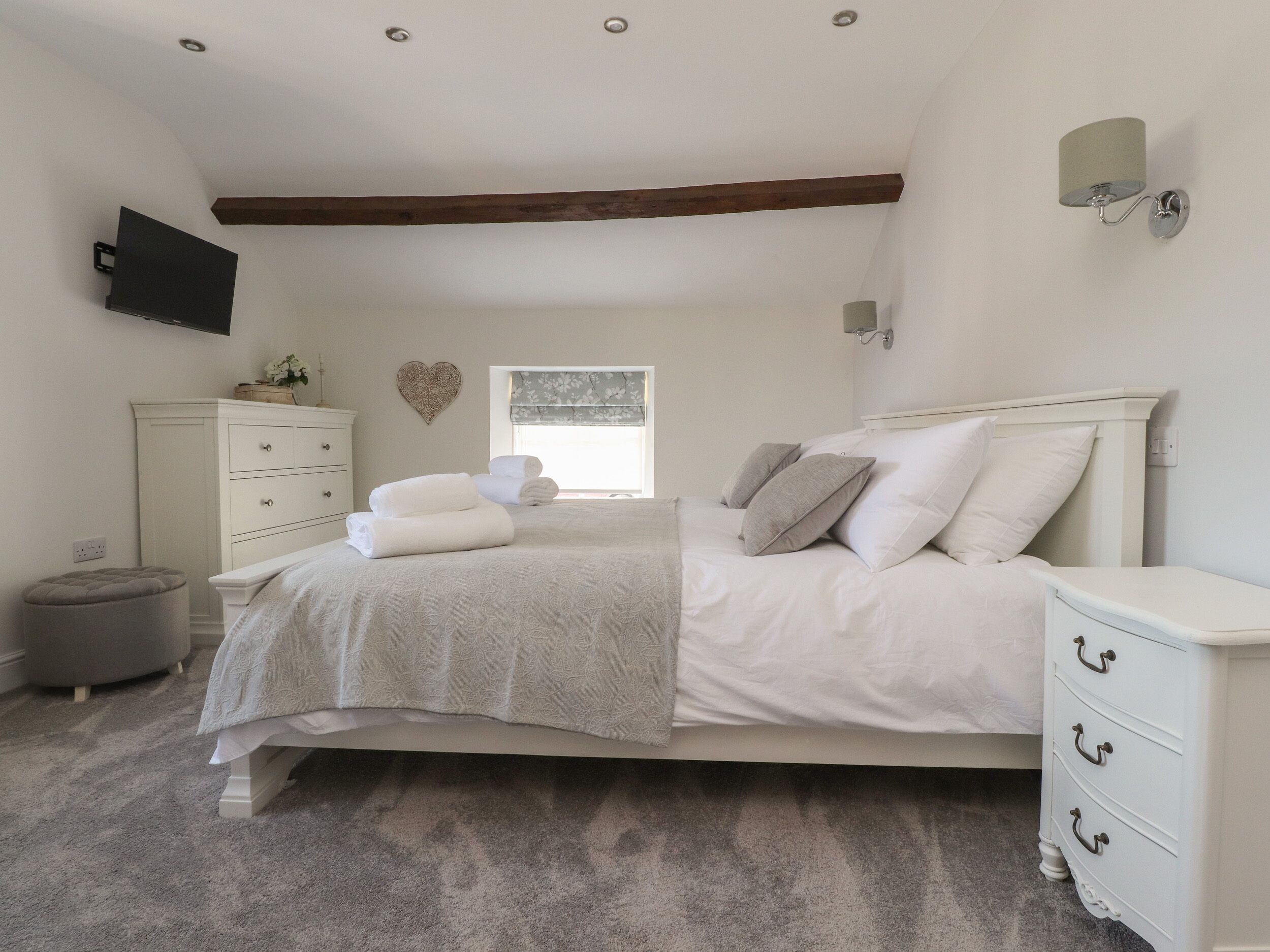 A bedroom with a bed, a dresser, a nightstand, a wall-mounted TV, a heart-shaped decoration, and a window with blinds.