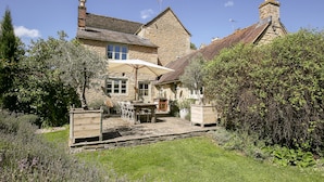 Une maison en pierre avec un toit de tuiles, un jardin où poussent des plants de lavande et un parasol blanc.
