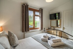 A bedroom with a bed, a window with curtains, a bedside table with a lamp, a dresser, and a television mounted above a mirror. - Lakeview Lodge (Carnforth)
