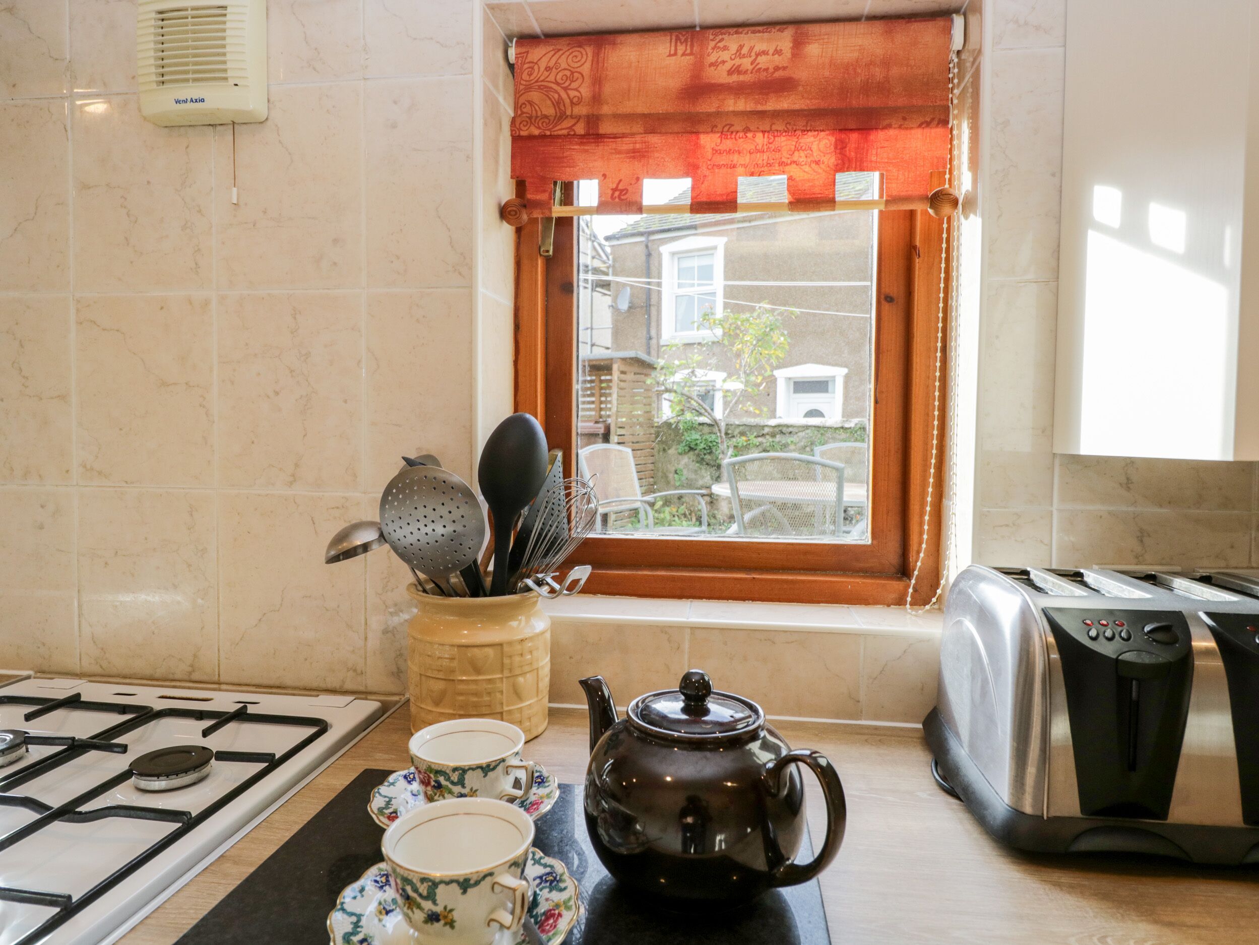 Una cocina con estufa a gas, hervidor, dos tazas de té, una ventana con vistas al exterior y un tostador.