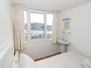A bedroom with a bed, a window with curtains, a view of a town and water, a small bathroom with a sink and mirror. - 21 Above Town (Dartmouth)