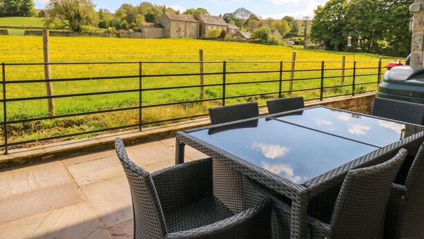 An outdoor patio with wicker furniture, a glass table, and a view of a green field and houses. - White Lea (Richmond)