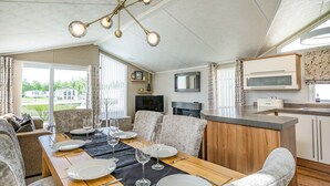 Ein Essbereich mit einem Holztisch, gedeckt für vier Personen, umgeben von gepolsterten Stühlen. Eine moderne Pendelleuchte hängt darüber, und ein Fernseher ist an der Wand montiert.