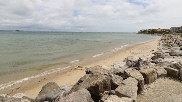 A beach with a rocky shoreline, calm water, and a distant view of boats and buildings.