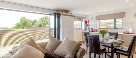 A modern living room with a sofa and chairs, a dining table with chairs, and a kitchen area with a window.