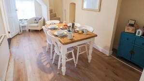 A dining area with a wooden table set for four, surrounded by white chairs. A clock and a framed mirror are on the walls.