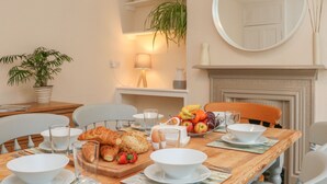 Un coin repas avec une table en bois mise pour le petit-déjeuner, comprenant des croissants, des fruits et des bols. Des chaises sont disposées autour de la table, ainsi qu'une plante en pot et un miroir accroché au mur.