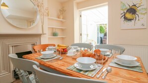 Une salle à manger avec une table en bois dressée pour quatre personnes, entourée de chaises blanches. On peut voir sur la table des bols, des assiettes et un saladier rempli de fruits. Un miroir est accroché au mur au-dessus de la table, et un tableau est visible sur le mur à droite.