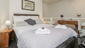 Een net opgemaakt bed met een gewatteerde deken, een nachtkastje met een lamp, een houten nachtkastje, een houten bankje, een spiegel en een geïnspireerd citaat in een lijst op de muur.