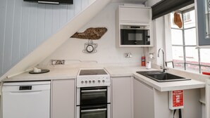 Een moderne keuken met een vaatwasser, kookplaat, oven en een ingebouwde magnetron. Onder het raam ligt een branddeken en aan de muur hangt een klok.
