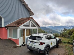 Un SUV Dacia blanc est garé devant une maison avec une porte rouge et un petit abri de jardin.