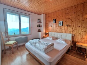 Un dormitorio con una cama grande, cabecera de madera, ventana con vistas, una silla y una mesita de noche con una lámpara.