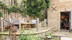 A stone building with a wooden deck, wicker furniture, and a garden area with various plants and flowers. - 1 Oakleys (Holmfirth)