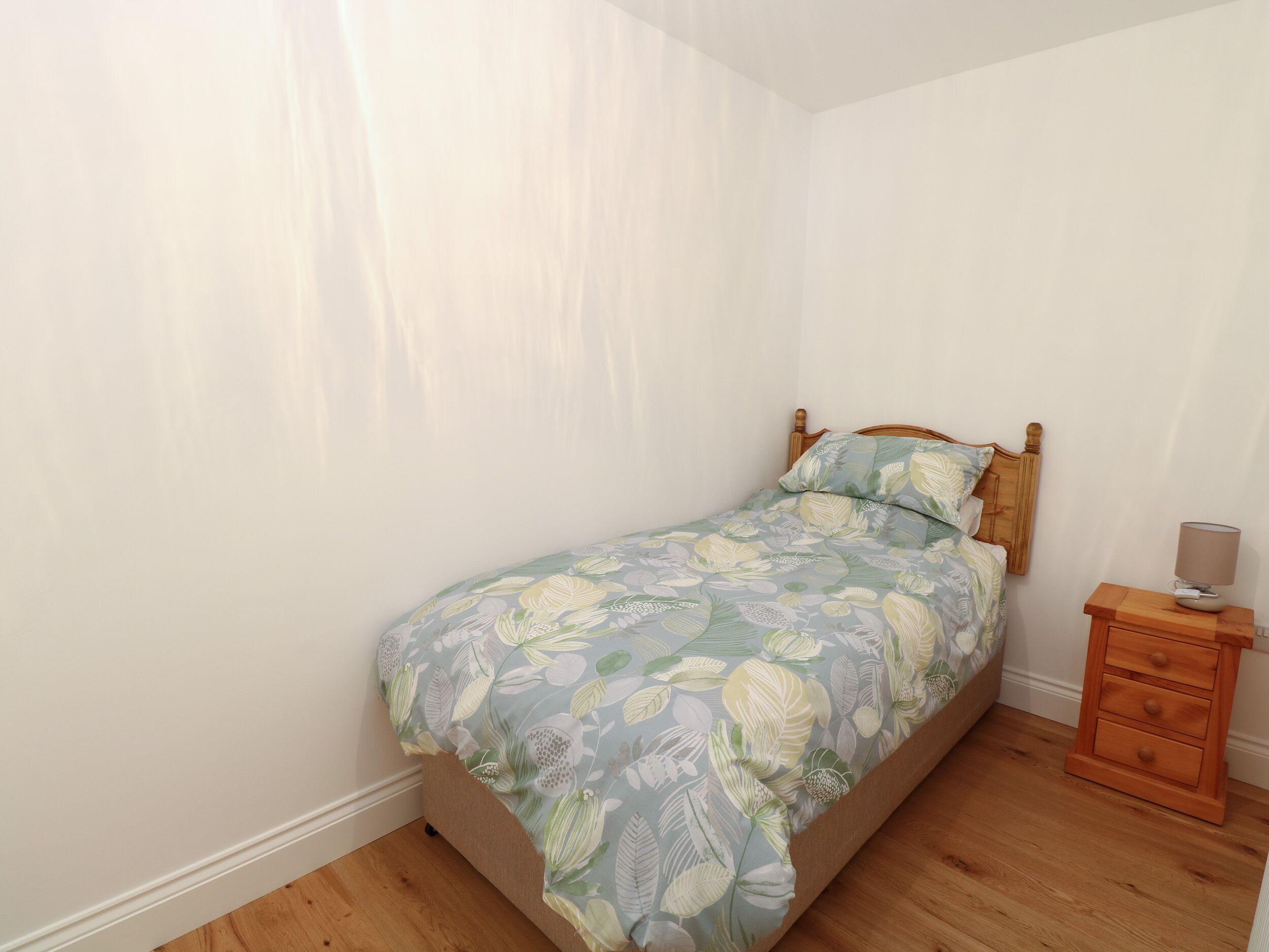 A bedroom with a single bed, a wooden nightstand with a lamp, and a plain white wall.