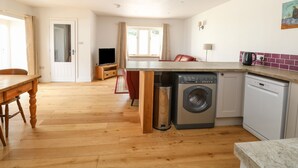 A compact kitchen with a washing machine, a dining area with a wooden table, and a TV in the background. - Waveney View Cottage (Great Yarmouth)