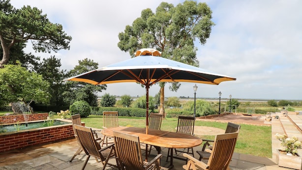 A patio with a wooden table and chairs, a large umbrella, and a view of a garden and distant landscape. - Waveney View Cottage (Great Yarmouth)