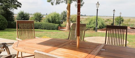 A wooden outdoor dining area with a table and chairs, surrounded by a garden with trees and a view of the countryside.