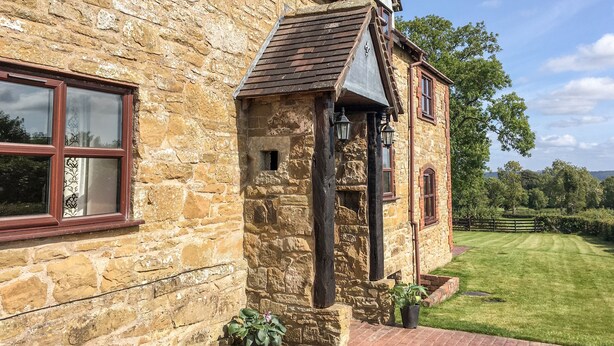 A stone building with a traditional design, featuring a window with a decorative frame, a small door, and a triangular roof structure. - Wilstone Cottage (Church Stretton)