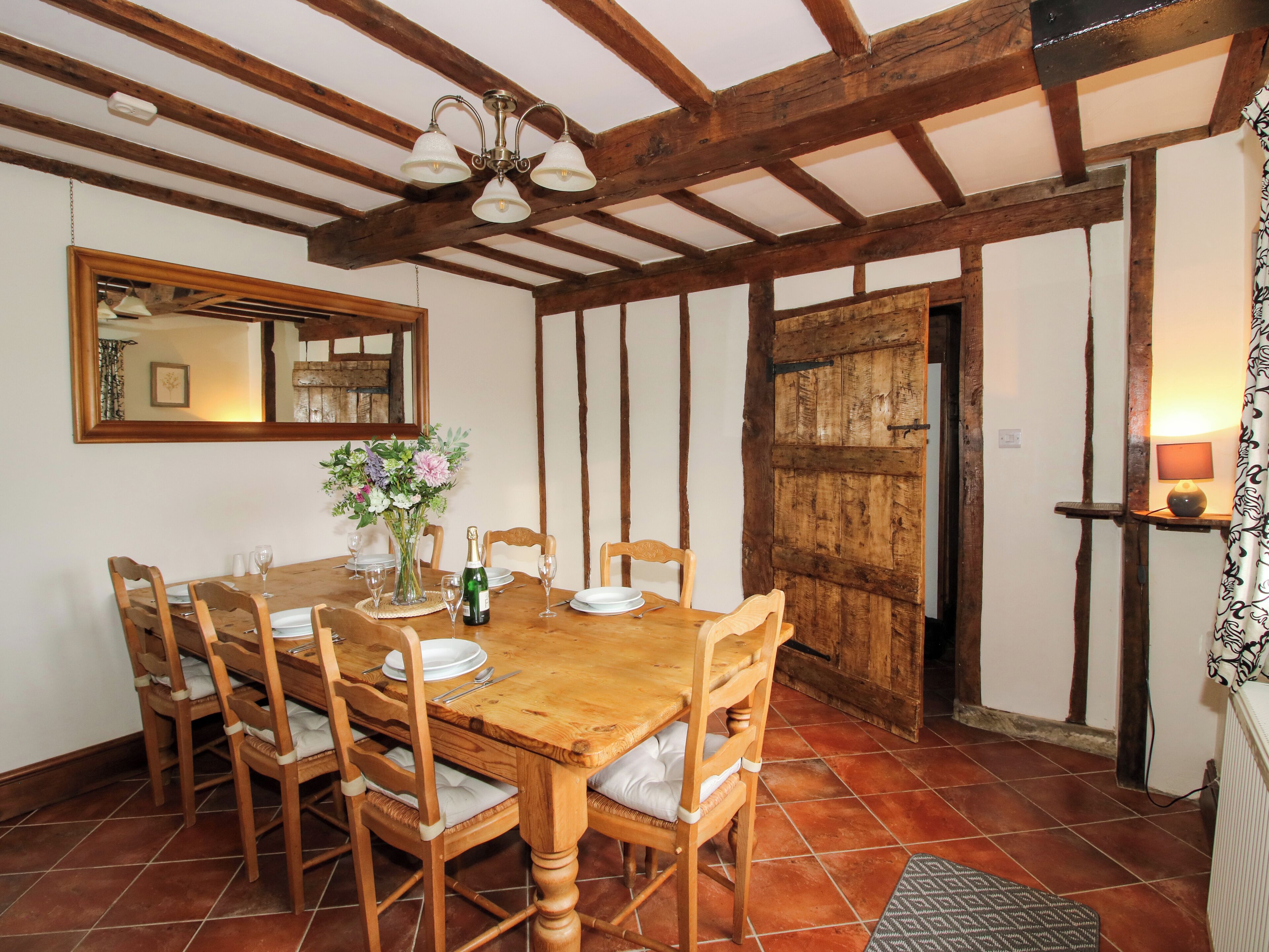 Een eethoek met een houten tafel voor zes personen, omringd door houten stoelen met beklede zitplaatsen. Een kroonluchter hangt aan het plafond, en een spiegel is aan de muur boven de tafel bevestigd.