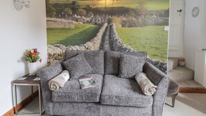 A grey sofa with rolled-up blankets, a small table with a vase of flowers, and a large scenic mural on the wall. - Town Head Cottage (Skipton)